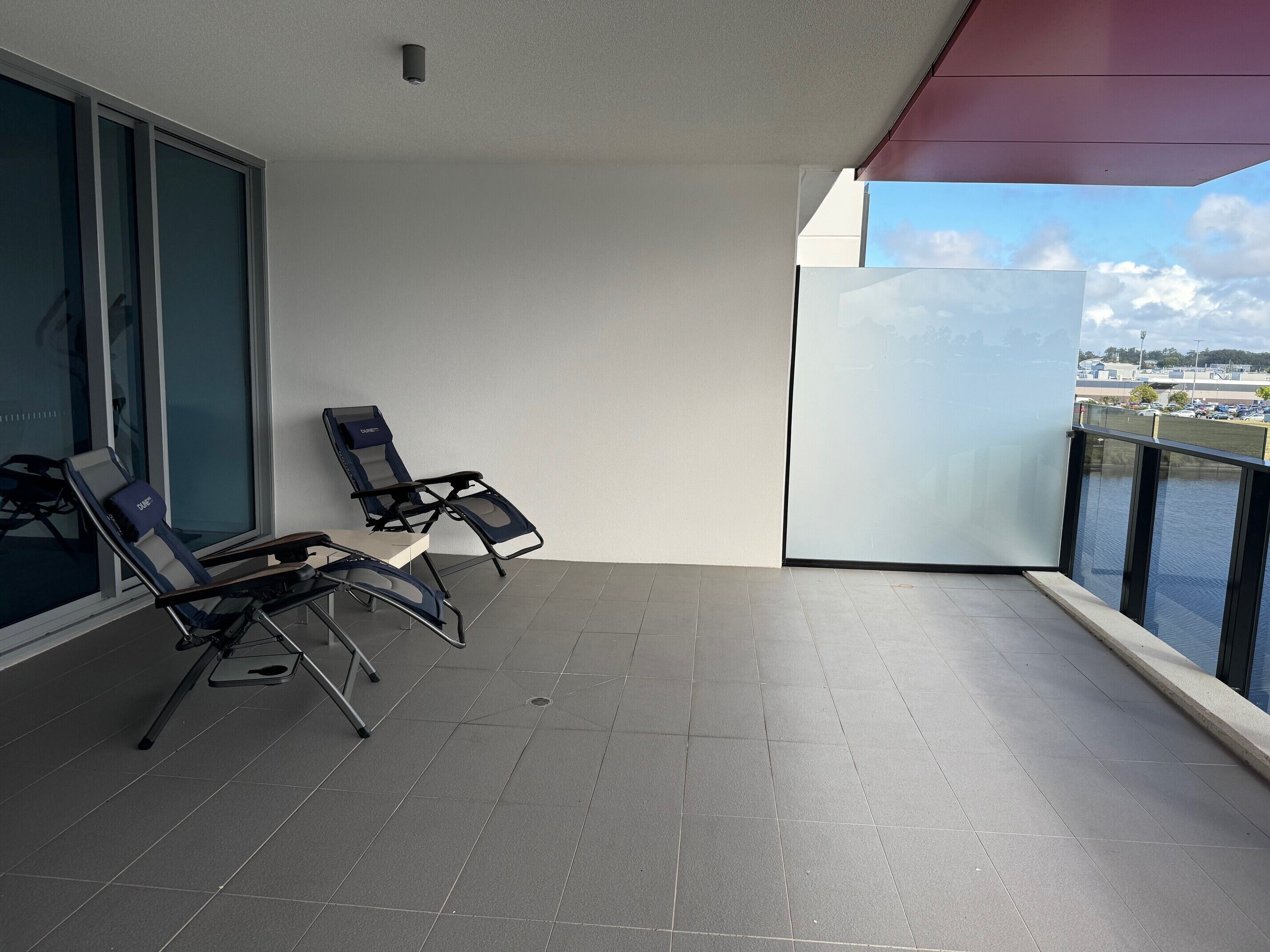 A balcony with two lounge chairs and a table, overlooking a body of water and a distant cityscape.