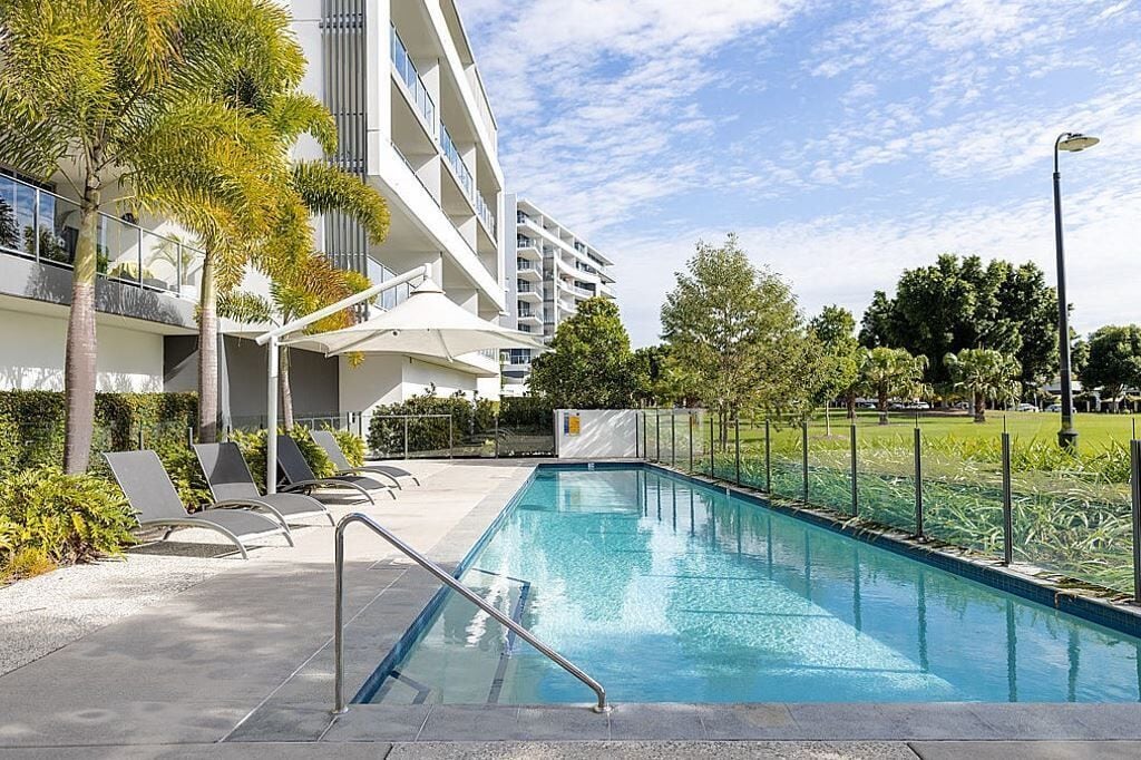 A swimming pool with lounge chairs, a covered seating area, and a modern building with balconies.