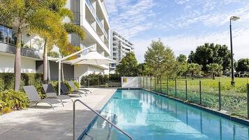 A swimming pool with lounge chairs, a covered seating area, and a modern building with balconies.