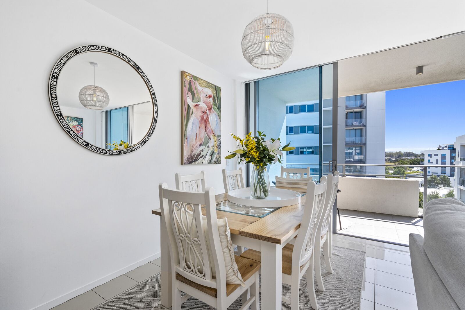 A modern dining area with a round table, white chairs, a vase with flowers, a large mirror, and a city view through large windows.