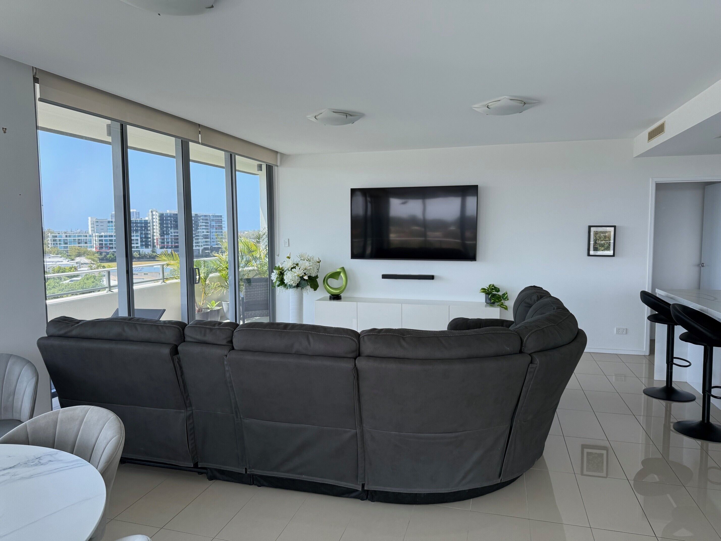 A modern living room with a large sectional sofa, a flat-screen TV mounted on the wall, and a dining area with a bar.