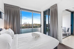 Habitación de hotel con una cama grande, vistas de la ciudad a través de ventanales del suelo al techo y un balcón con mesa y sillas.