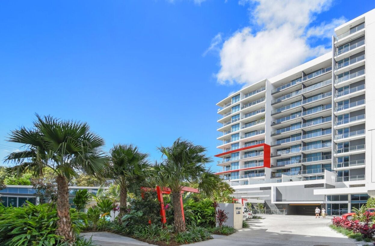 Een modern hotel met meerdere verdiepingen, een verzorgde tuin met palmbomen en een helderblauwe lucht.