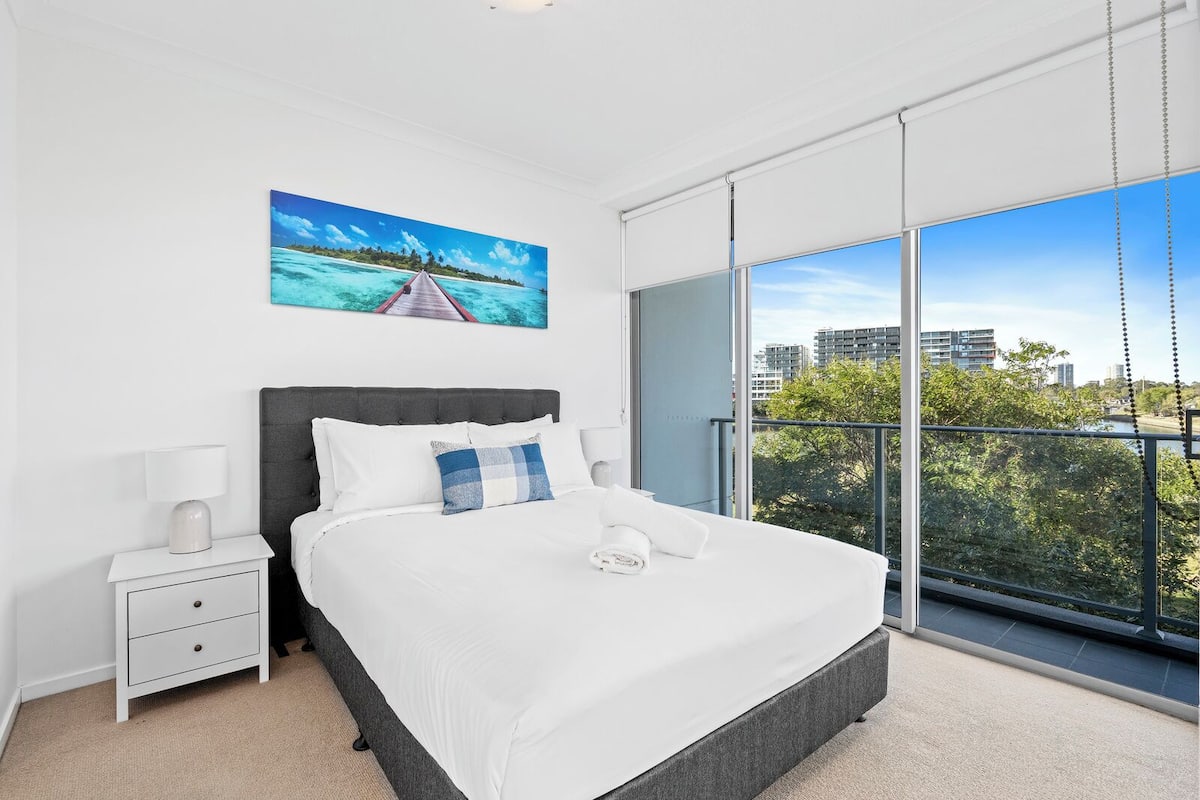 Ein modernes Schlafzimmer mit einem großen Bett, einem Nachttisch, einem Bild von einem tropischen Strand und einem Balkon mit Blick auf die Stadt.