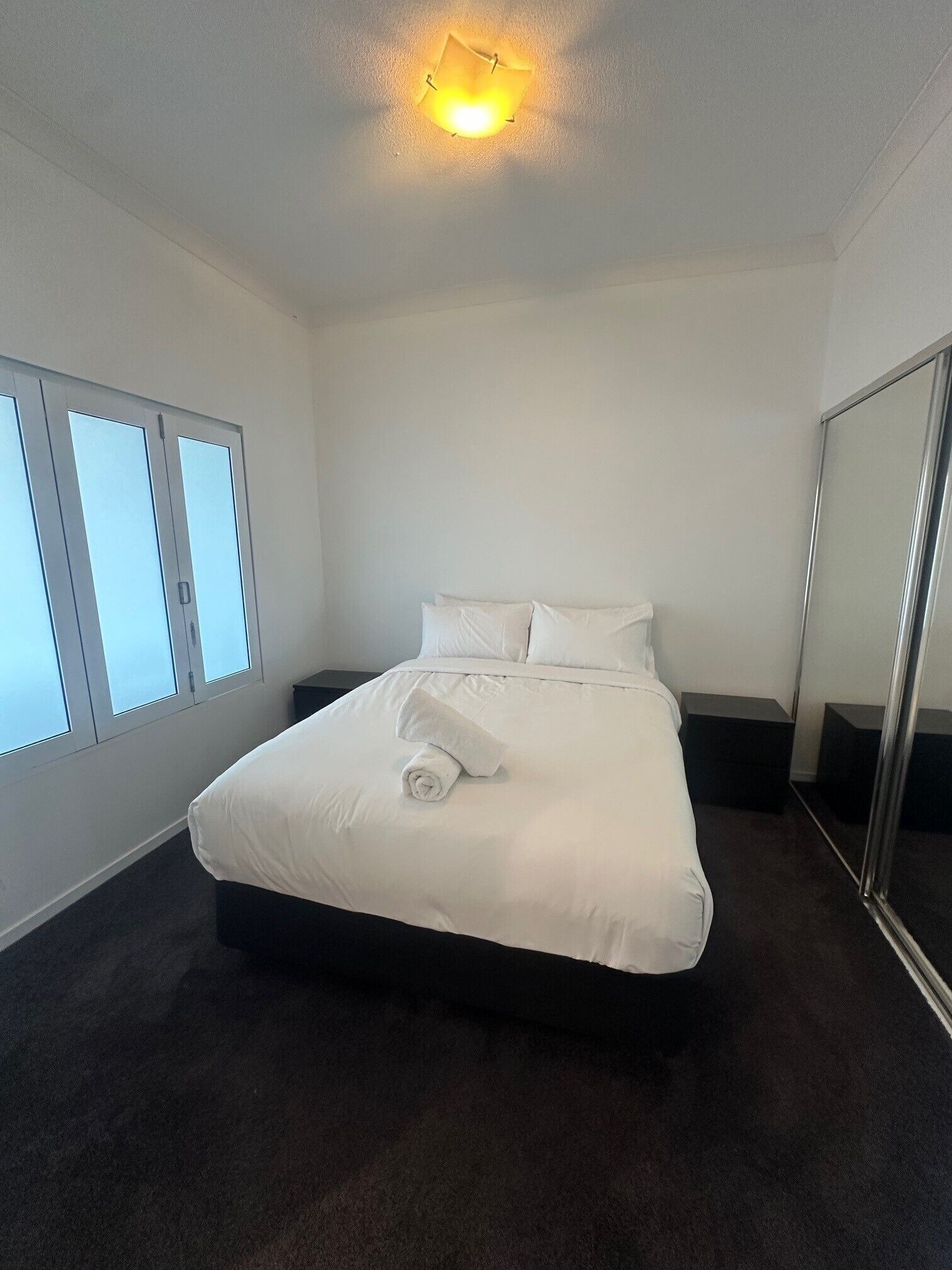 Ein ordentlich gemachtes Bett mit weißer Bettwäsche, ein Nachttisch, ein großer Spiegel und ein Fenster mit Jalousien.
