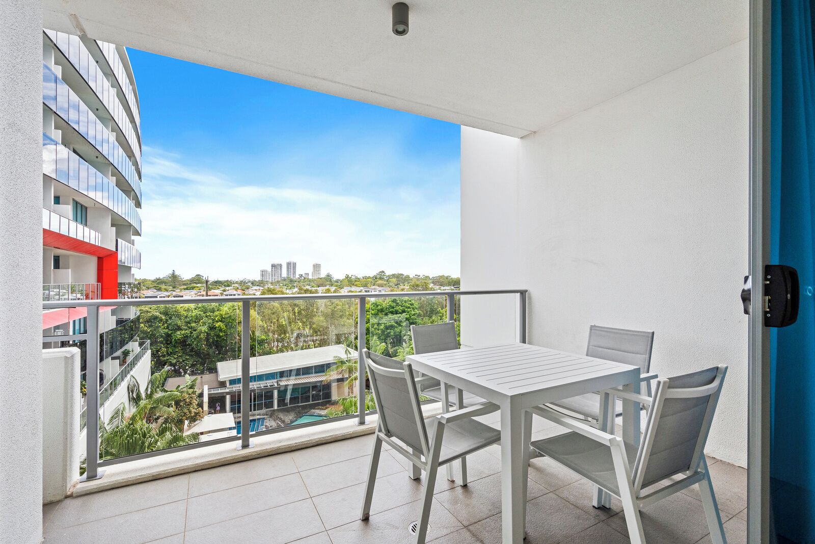 A balcony with a white table and chairs, a view of a cityscape, and a pool in the background.