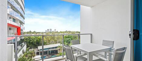 A balcony with a white table and chairs, a view of a cityscape, and a pool in the background.