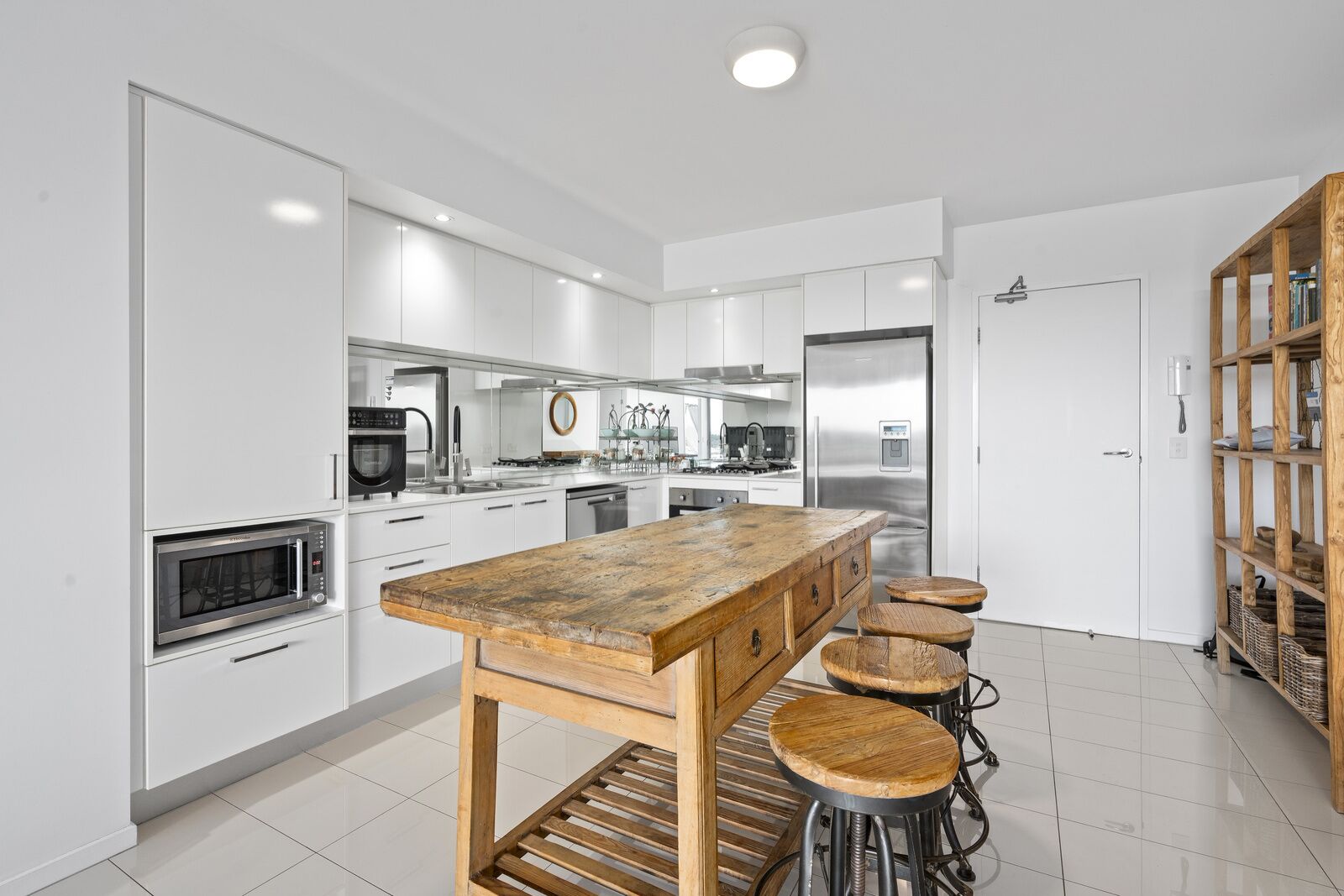 A modern kitchen with white cabinetry, stainless steel appliances, a wooden island with stools, and a built-in bookshelf.