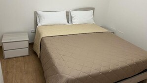 A single bed with a beige quilted bedspread and two white pillows, a small bedside table with drawers, and a plain white wall. - A1 SOLE - APARTMENT WITH 6 BEDS (Balestrate)
