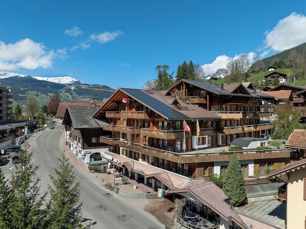 A multi-story wooden building with balconies, a ski resort in the background, and a road in the foreground. - Cosy apartment for 2 guests with WIFI, TV and pets allowed (Grindelwald)