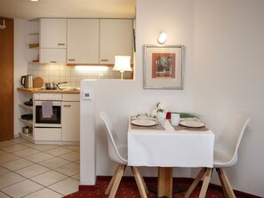 Uma cozinha compacta com armários brancos, fogão e geladeira. Há uma mesa de jantar com cadeiras e um quadro na parede.