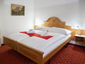 Una cama de madera con un cojín rojo decorativo, ropa de cama blanca y una mesita de noche con una lámpara.