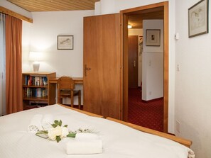 Quarto de hotel com cama, escrivaninha, estante e porta que dá para outro cômodo.