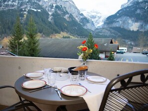 Mesa para dos personas con platos, cubiertos, vasos y un jarrón con flores, con vistas a un paisaje montañoso.