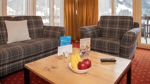 Une chambre d’hôtel avec deux canapés à carreaux, une table basse en bois sur laquelle sont disposés des fruits et une télécommande, et une vue sur des montagnes enneigées par de grandes fenêtres.