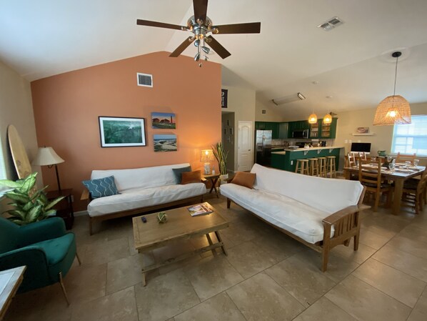 A living room with two sofas, a coffee table, and a dining area with a table and chairs. There is a ceiling fan, a kitchen in the background, and a plant on the left. - Keep it Shrimple- short walk to beach, pool, pets friendly! (Port Aransas)