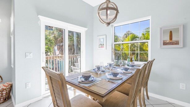 A dining area with a long table set for six, white walls, and large windows offering a view of palm trees. - La Coconut: 1 Block to Beach! Dog Friendly! Ping Pong & Foosball, Heated Pool by Anna Maria Life (Holmes Beach)