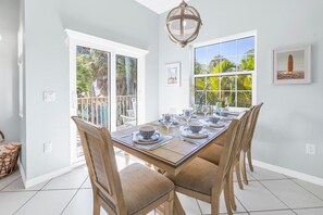 Un comedor con una mesa larga dispuesta para seis personas, paredes blancas y ventanas amplias con vistas a palmeras.