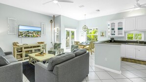 A modern living room with a grey sofa, a wooden coffee table, and a TV. There is a kitchen with white cabinets and a dining area with a table and chairs. - La Coconut: 1 Block to Beach! Dog Friendly! Ping Pong & Foosball, Heated Pool by Anna Maria Life (Holmes Beach)