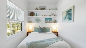 A bedroom with a bed, two bedside tables, a window with blinds, a shelf with decorative items, and a framed picture on the wall. - La Coconut: 1 Block to Beach! Dog Friendly! Ping Pong & Foosball, Heated Pool by Anna Maria Life (Holmes Beach)