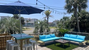 A wooden deck with a blue sofa, white stools, a table, and a large umbrella.