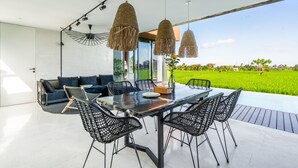 An outdoor dining area with a table and chairs, hanging lights, and a view of a green field. - Green Flow Villa 17 (Ubud)