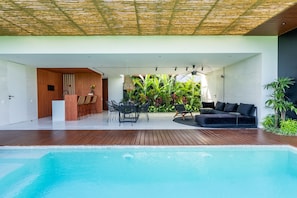 Ein moderner Poolbereich im Freien mit einer Holzterrasse, einer Lounge mit einer schwarzen Couch und Sesseln sowie einer üppigen, grünen Pflanzenwand im Hintergrund.