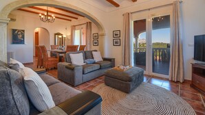 A spacious living room with a grey sofa set, a wooden coffee table, and a patterned rug. There is a dining area with wicker furniture and a chandelier. A large sliding glass door leads to an outdoor balcony with a view of a mountain.