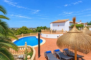 A pool area with a white building, lounge chairs, and a thatched umbrella.