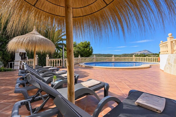 A poolside area with lounge chairs, a thatched umbrella, and a view of a mountain.