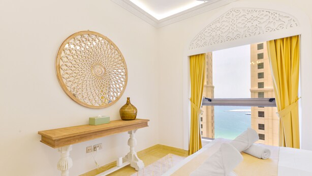 A hotel room with a bed, a wooden console table, a decorative wall piece, and a view of the sea through a window with yellow curtains.