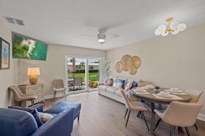 A modern living room with a dining area, a ceiling fan, and a large window overlooking a garden.