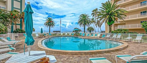 A pool area with lounge chairs and umbrellas, surrounded by palm trees and a building.
