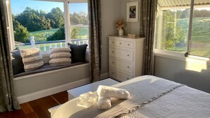Een slaapkamer met een groot bed, een raamzitje met kussens, een kaptafel en uitzicht op een groen landschap.
