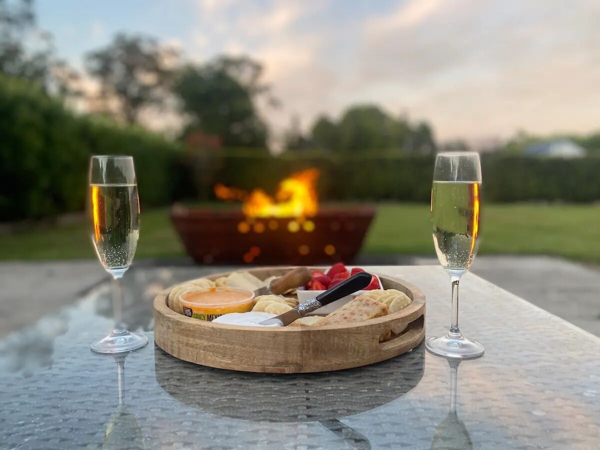 Eine runde Holzplatte mit Käse, Crackern und einem Messer auf einem Tisch, auf dem zwei Gläser Weißwein stehen. Im Hintergrund ist eine Feuerstelle zu sehen.