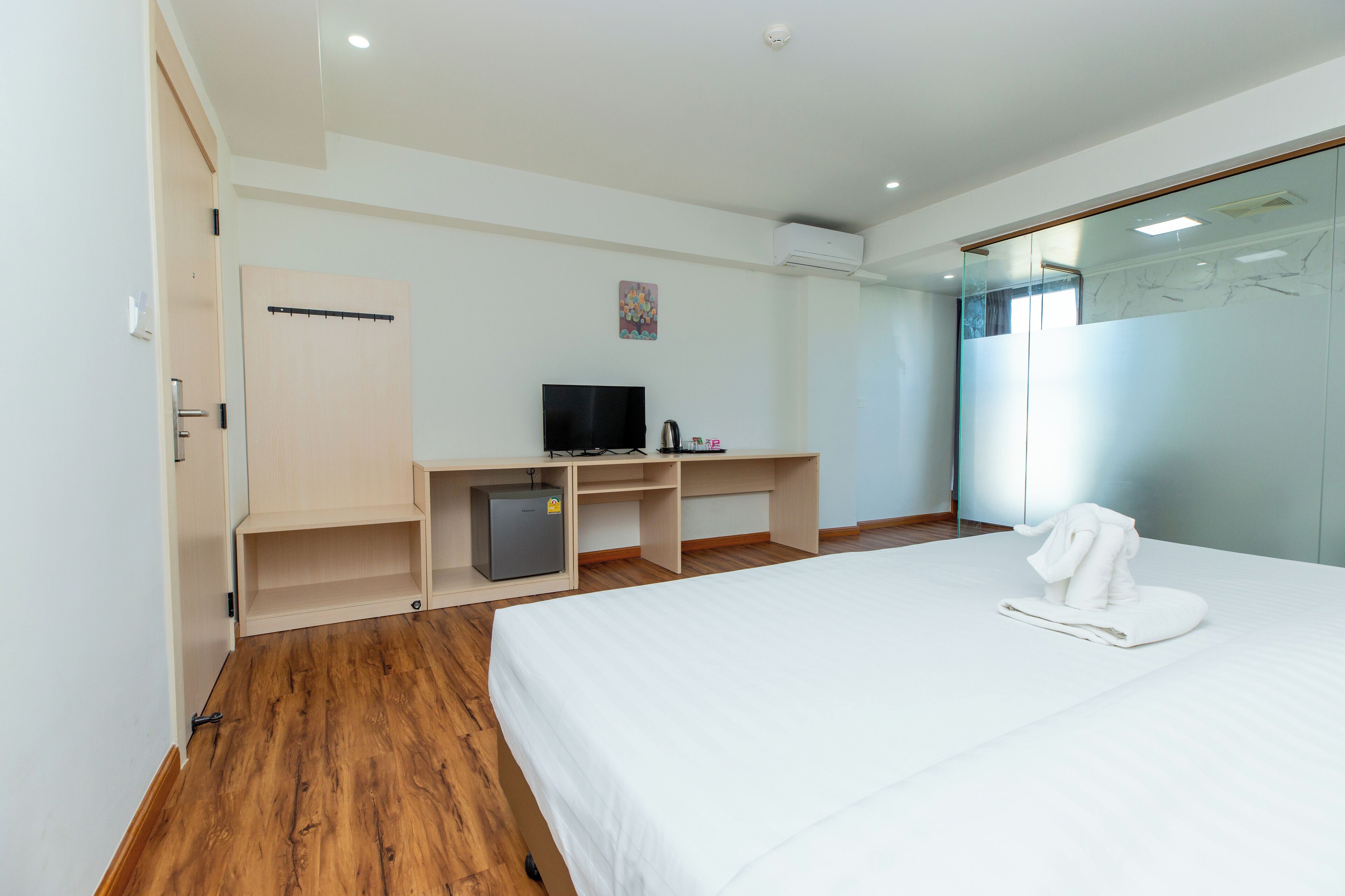 Una camera d'albergo moderna con pavimento in legno, un letto con copriletto bianco, una scrivania con televisore e un piccolo frigorifero.