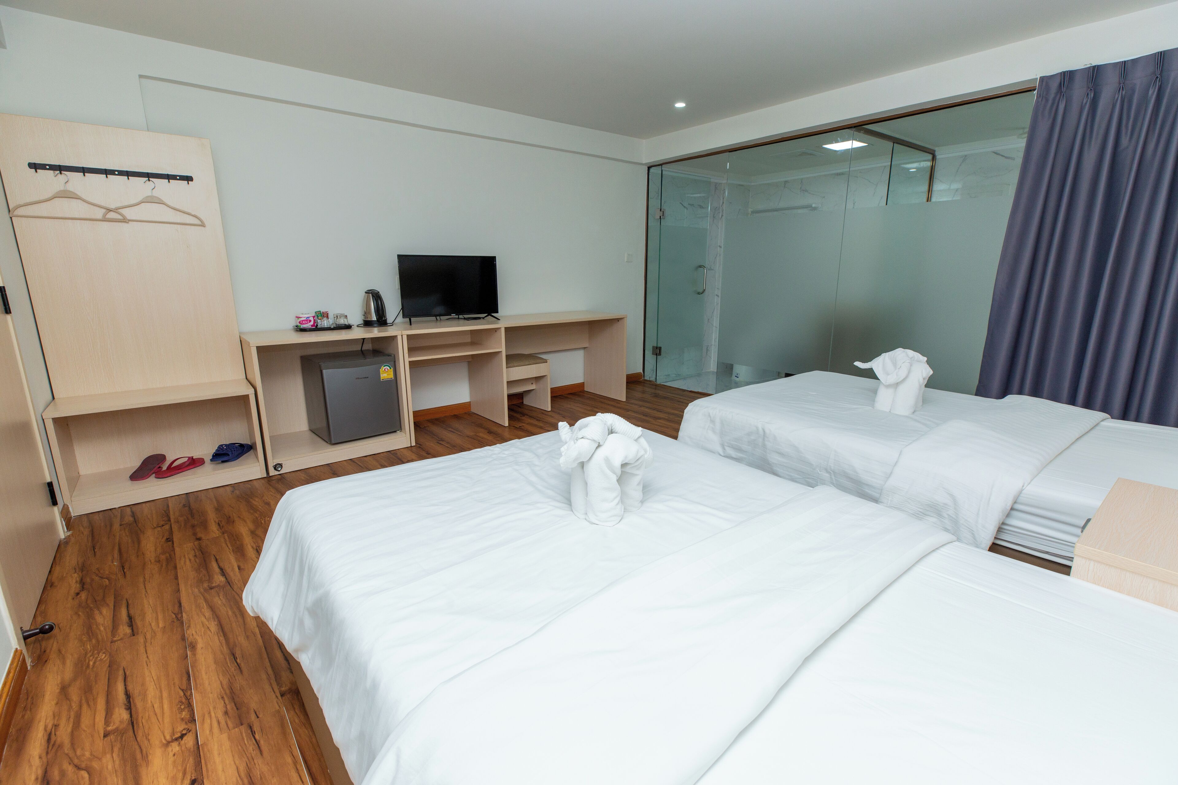 A hotel room with two beds, a desk with a TV, a wardrobe, and a shower area.