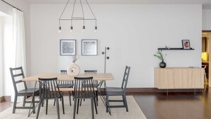 Et moderne spiseområde med et træbord, sorte stole, en hængende lampe og minimalistisk indretning med indrammede kunstværker og en lille plante.