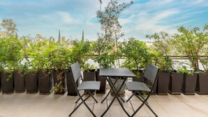 En udendørs terrasse med et bord og to stole, omgivet af krukker med planter og træer.