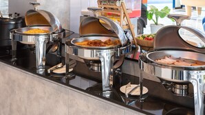 Buffet montado com diversos alimentos em rechauds metálicos, suporte de madeira com condimentos e uma tigela de frutas.