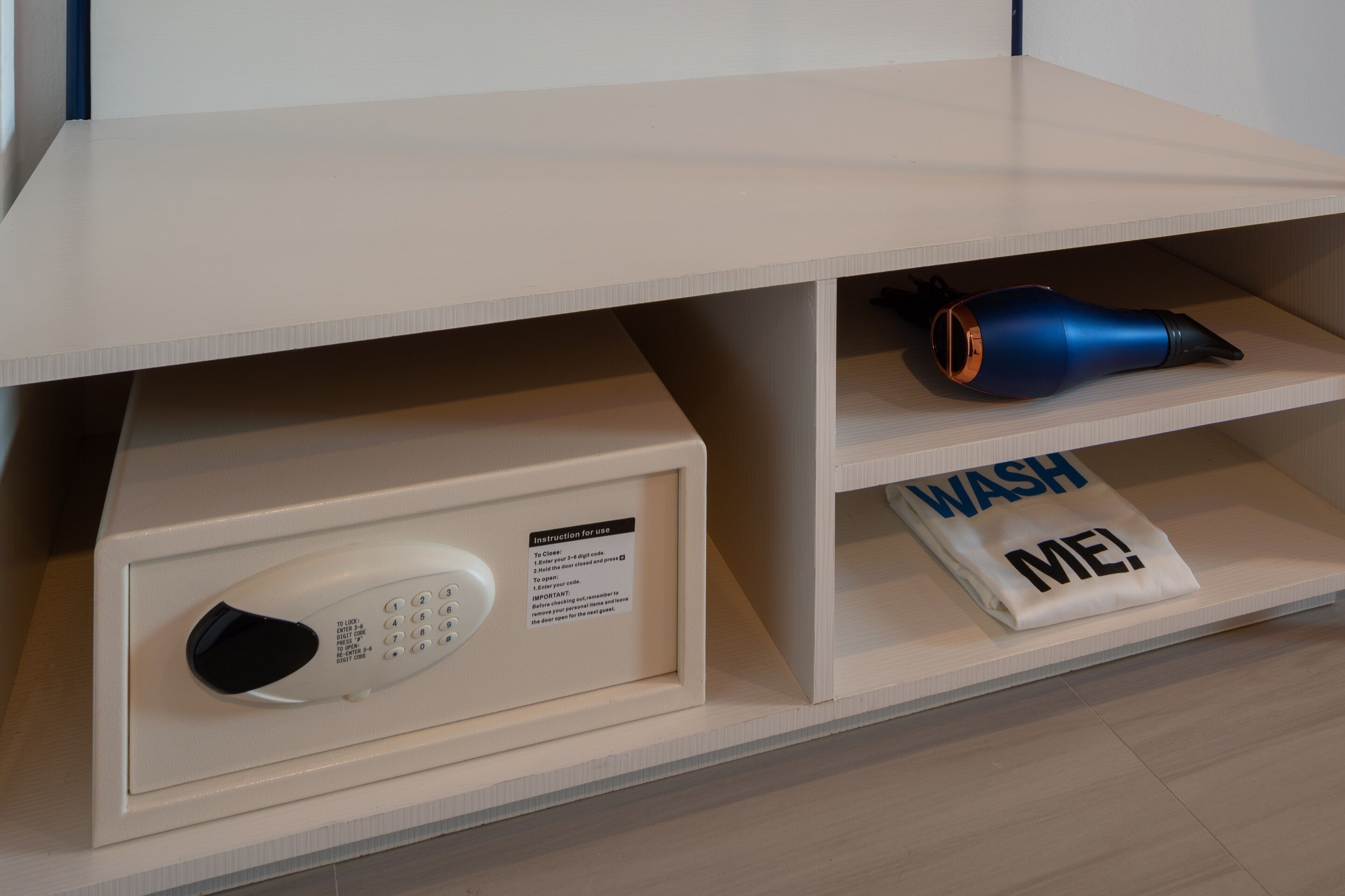 A microwave with a control panel and instruction label, a blue appliance on a shelf, and a towel with "WASH ME!" printed on it.