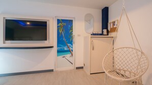 Quarto com televisão, porta decorada com um mural de praia, cadeira suspensa e pequeno armário.