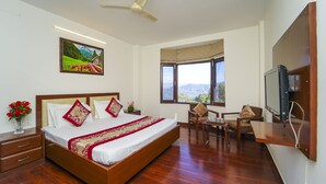 A hotel room with a large bed, a desk, a chair, a television, a painting, and a window with a mountain view. - Kamlia 3 BHK Villa By Dumnu Homes (Kasauli)