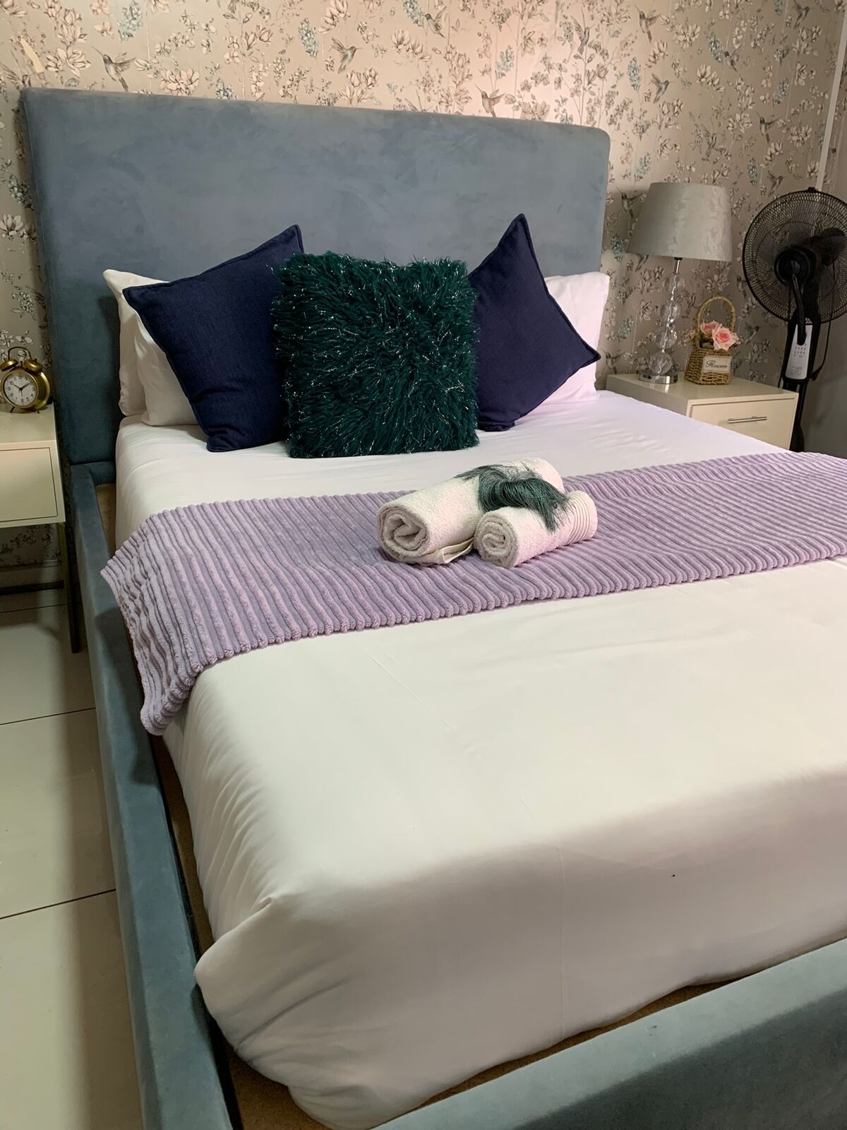 Una cama bien hecha con una colcha a rayas, toallas enrolladas, un cabecero azul y un papel tapiz floral.