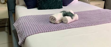 Una cama bien hecha con una colcha a rayas, toallas enrolladas, un cabecero azul y un papel tapiz floral.