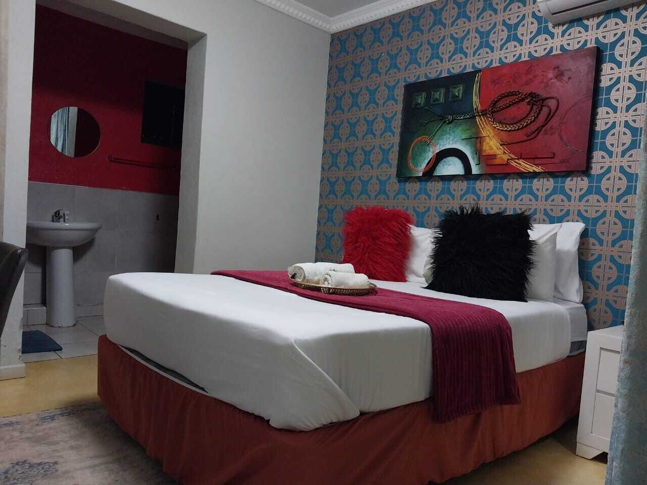 A bedroom with a bed, a red and white bedspread, a red throw blanket, and a black pillow. There is a painting on the wall and a sink in the corner.