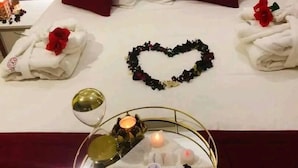 Una cama bien hecha con sábanas blancas, almohadas rojas y una decoración con forma de corazón hecha de flores. Hay una bandeja con una vela y un vaso de agua sobre la cama.