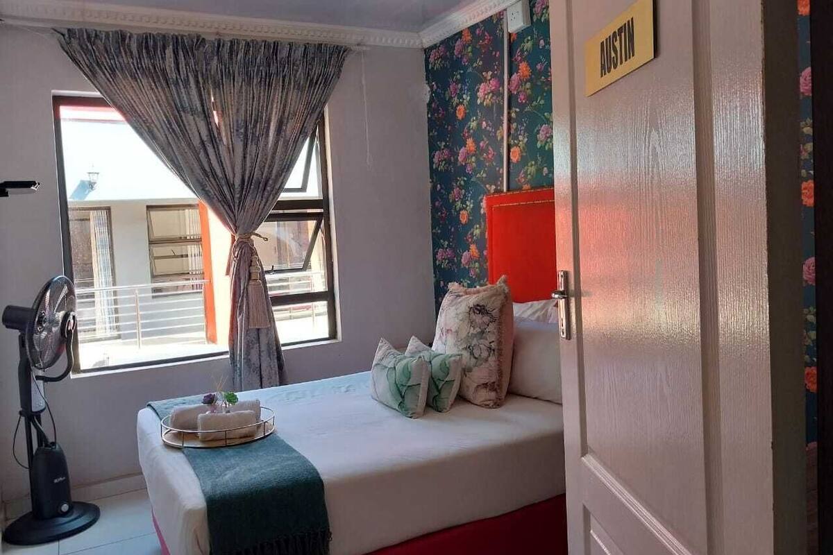 A room with a bed, a fan, a window with curtains, a floral wallpaper, and a door with a nameplate.