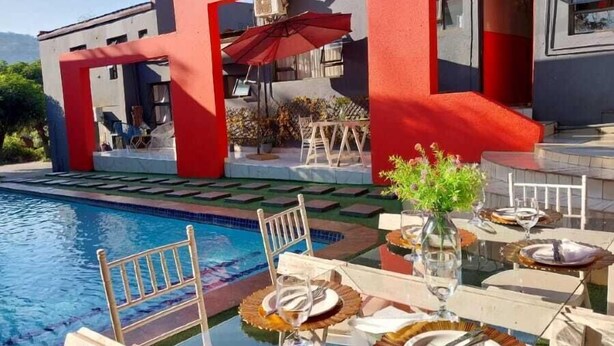 Un área de comedor junto a la piscina, con una sombrilla roja, asientos al aire libre y vistas a un edificio con columnas rojas.