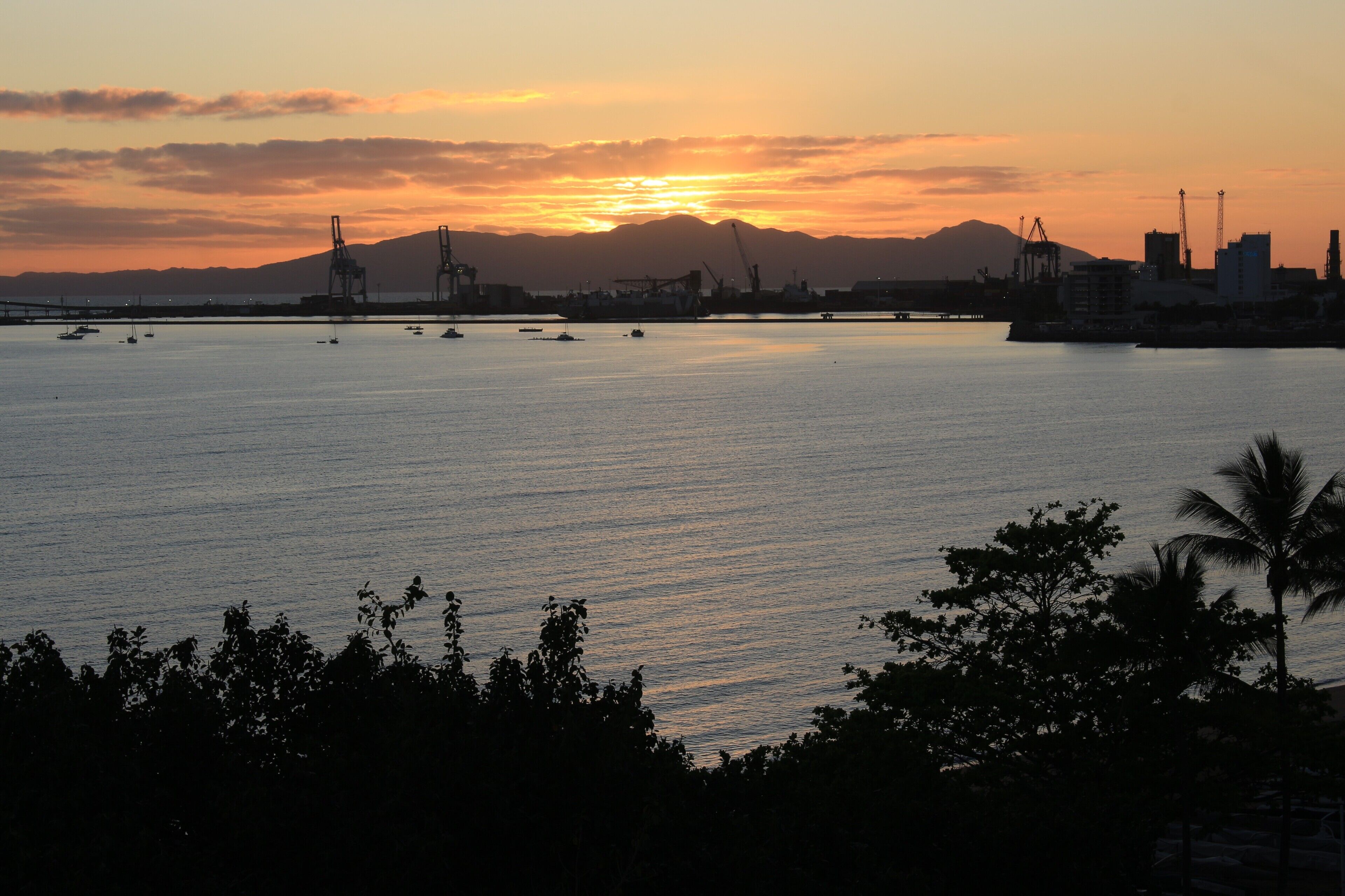 Eine Küstenstadtlandschaft bei Sonnenuntergang mit Booten auf dem Wasser, Industriegebäuden und einem Berg im Hintergrund.